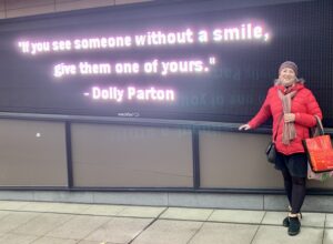 Pam in front of sign that reads "If you see someone without a smile, give them one of yours." - Dolly Parton